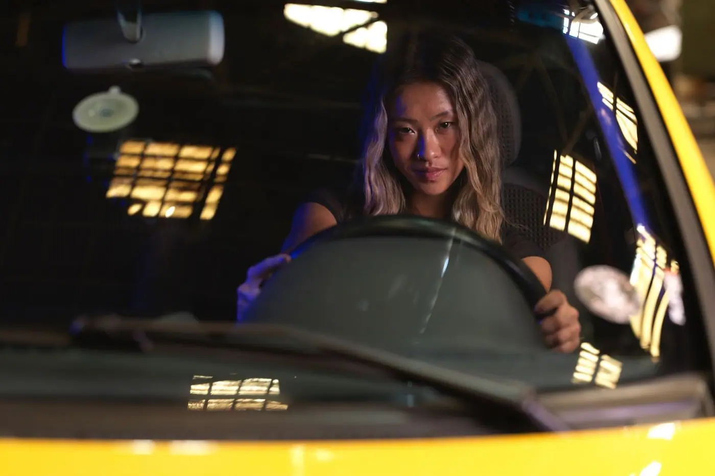 A woman confidently driving a yellow car during nighttime, cityscape reflections on windshield.
