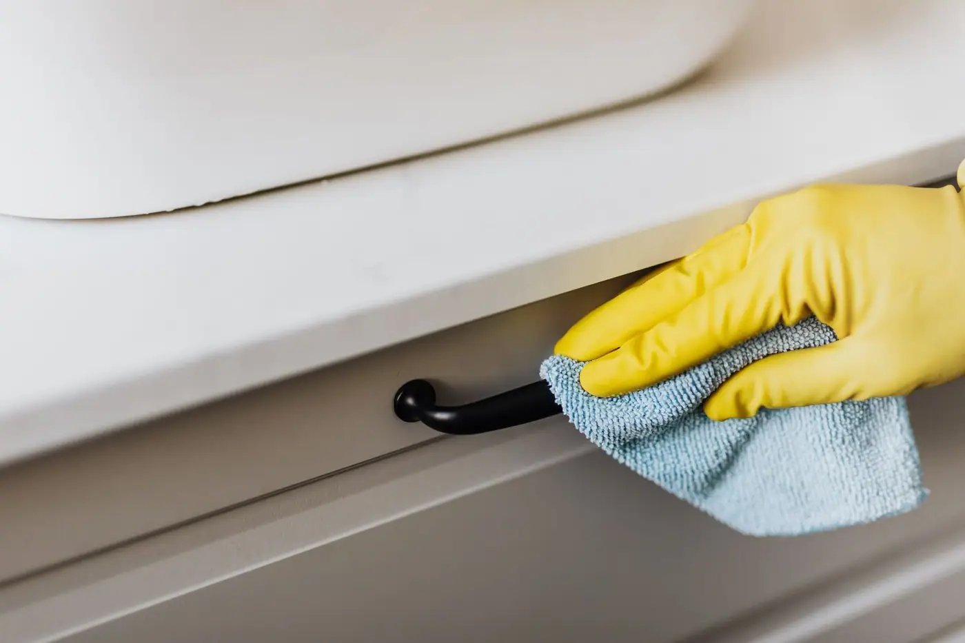Gloved hand cleaning a cabinet handle with a microfibre cloth in a bright bathroom
