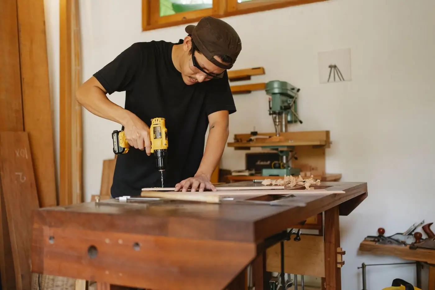 A man in a cap drilling a hole in a wooden plank at a workbench