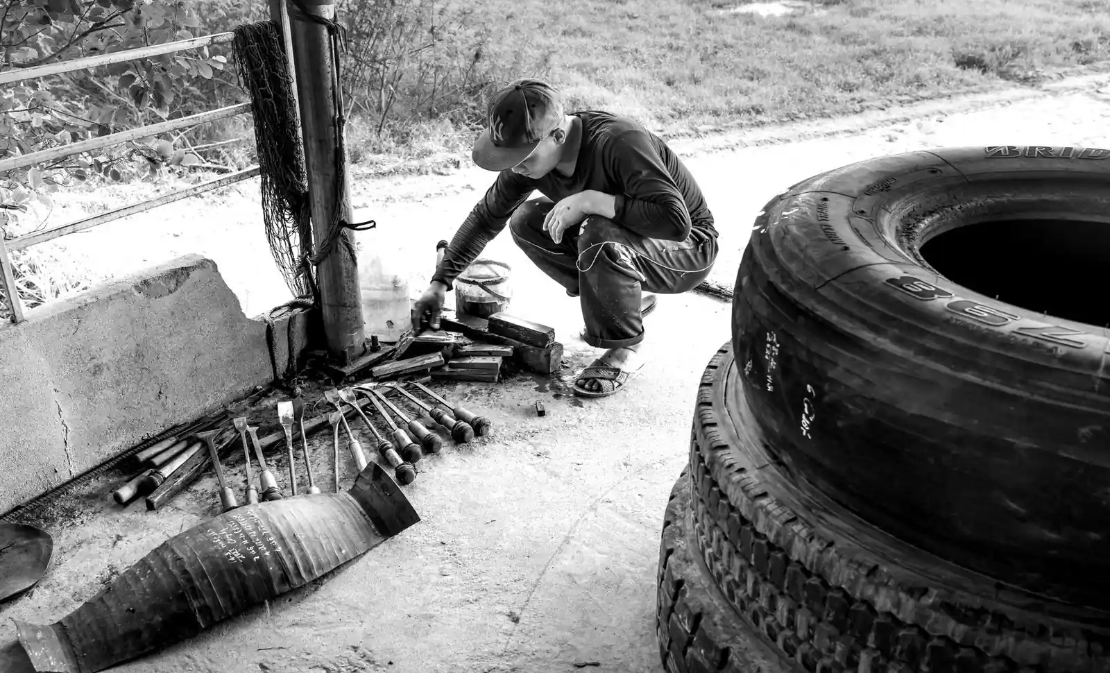 Black and white photo of a crouching worker with tools and tires in an outdoor setting