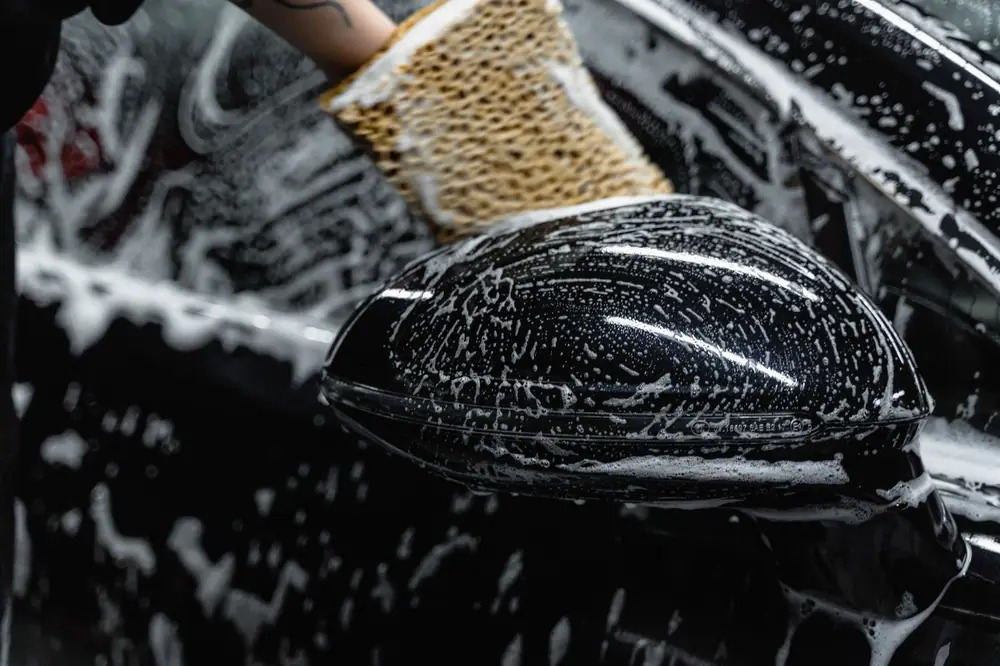 Hand scrubbing a car mirror with a sponge covered in soap at a car wash.