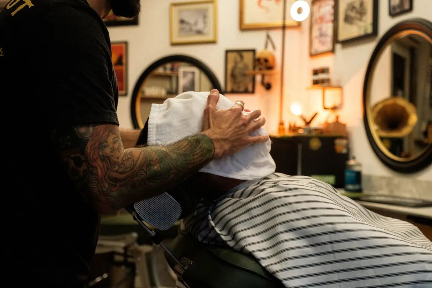 A barber with tattoo sleeves applying a hot towel in a vintage-style barbershop
