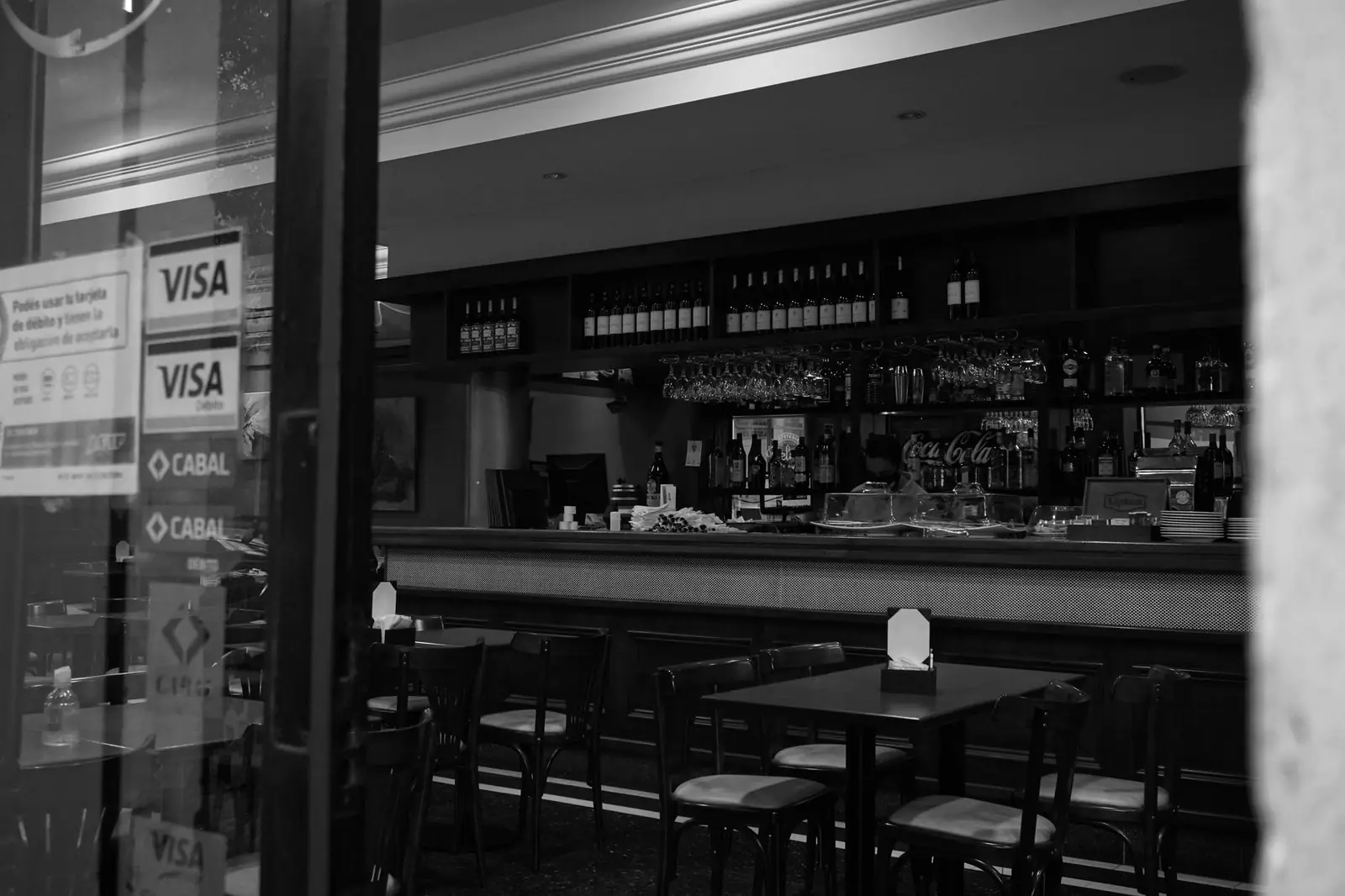 A black and white view of a cozy bar interior with empty seating and a well-stocked counter.