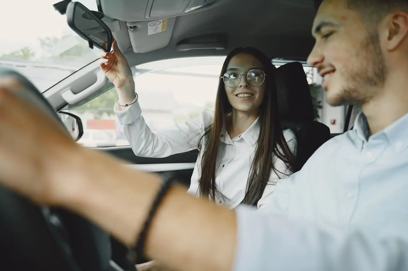 A cheerful young couple driving, the woman adjusting the mirror.