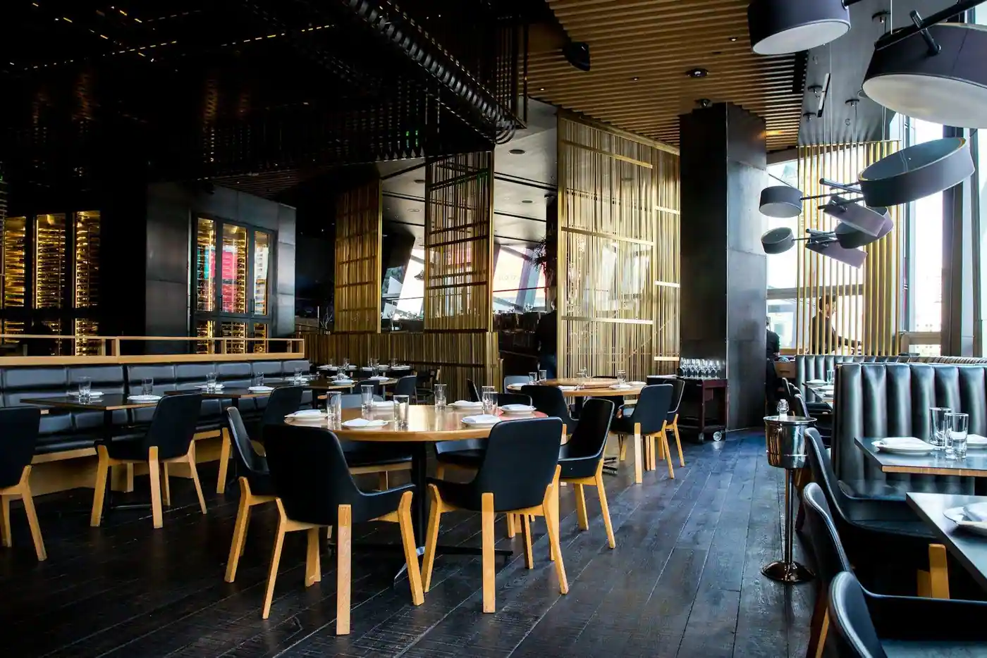 Restaurant interior with round wooden tables, black leather booths, wooden chairs and a glass-enclosed wine display