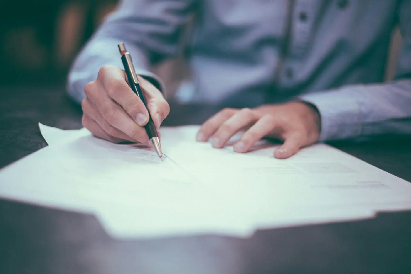 Close-up of a person in a light blue shirt signing documents with a pen at a desk.