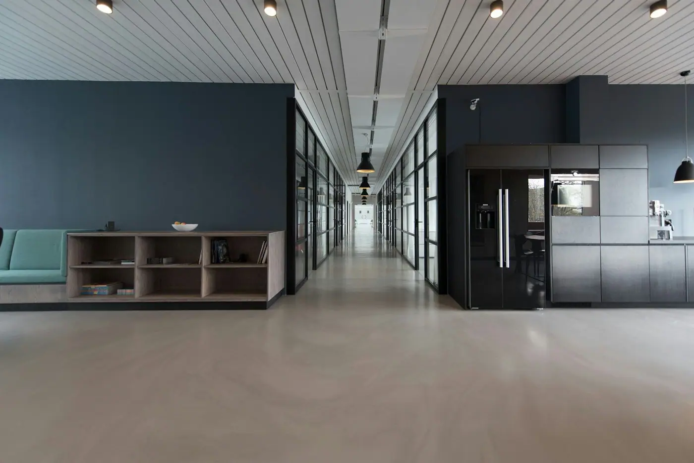 A spacious modern office interior with polished concrete floors, a long glazed corridor, built-in wooden shelving and a seating bench.