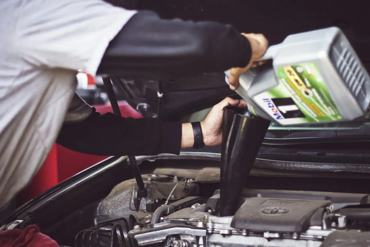 A mechanic pouring motor oil through a black funnel into a car engine under an open bonnet
