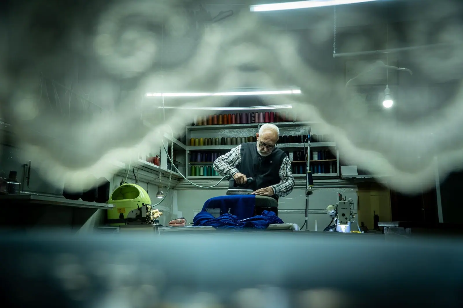 An elderly tailor meticulously working on fabric in a cozy workshop setting.
