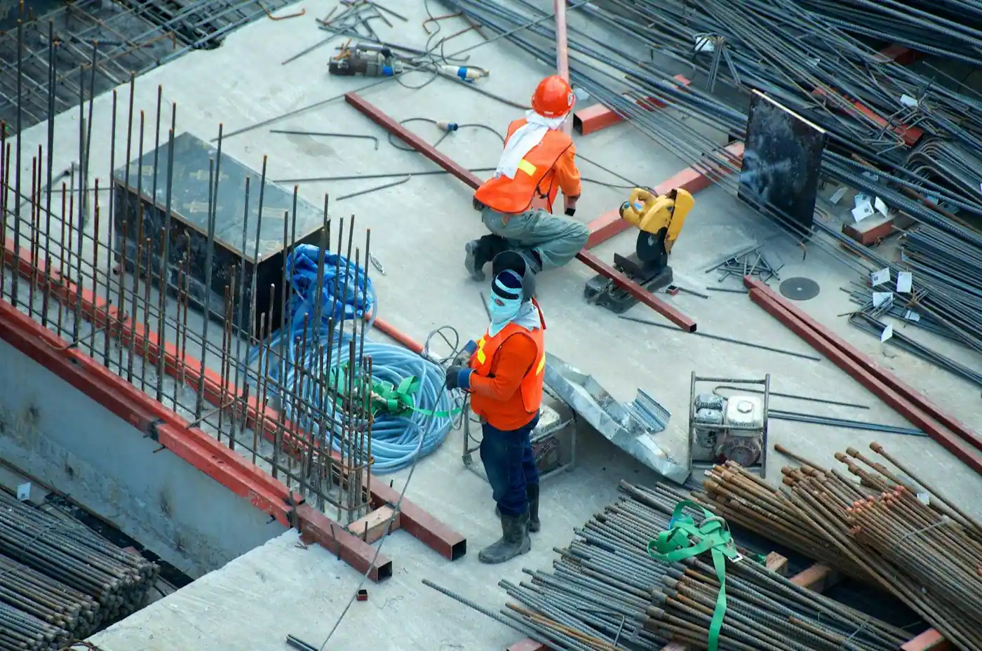 Overhead view of two construction workers in orange safety vests and hard hats working amid rebar, cables and equipment on a building site