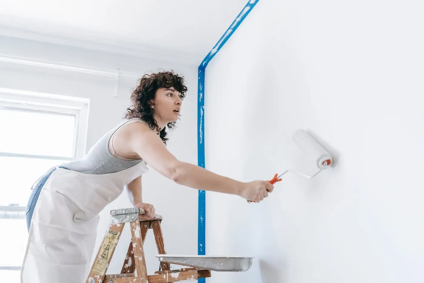 A woman on a ladder painting a wall white during a home renovation.
