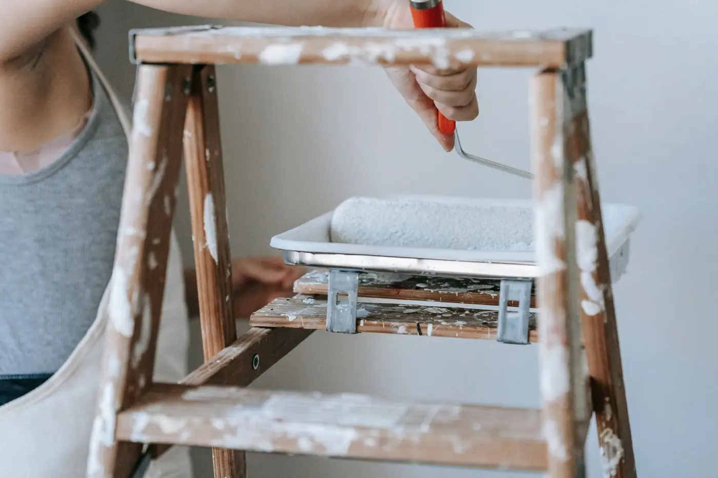 A woman using a roller and ladder to paint a room wall indoors.