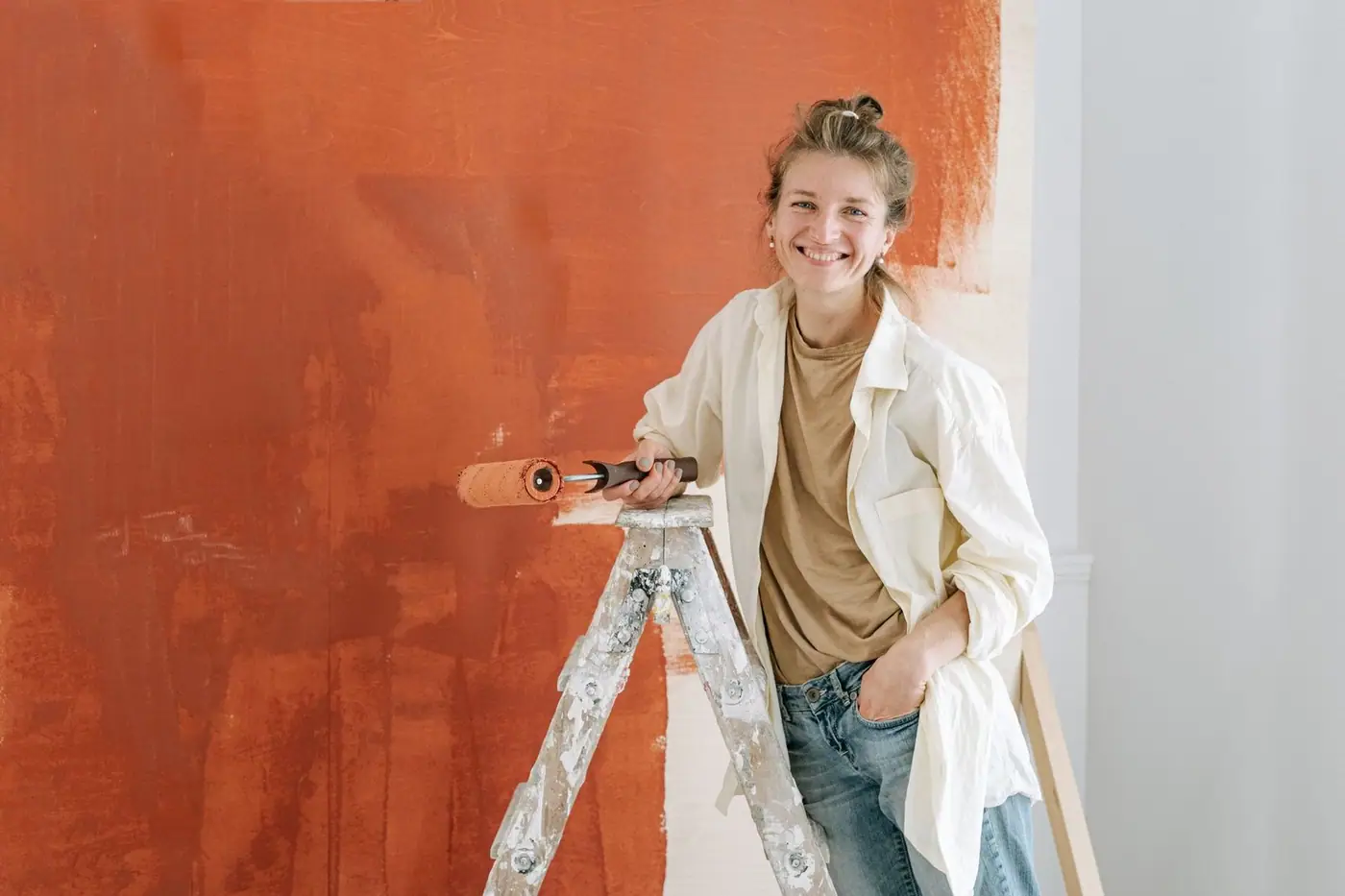 A woman on a ladder painting an orange wall indoors.