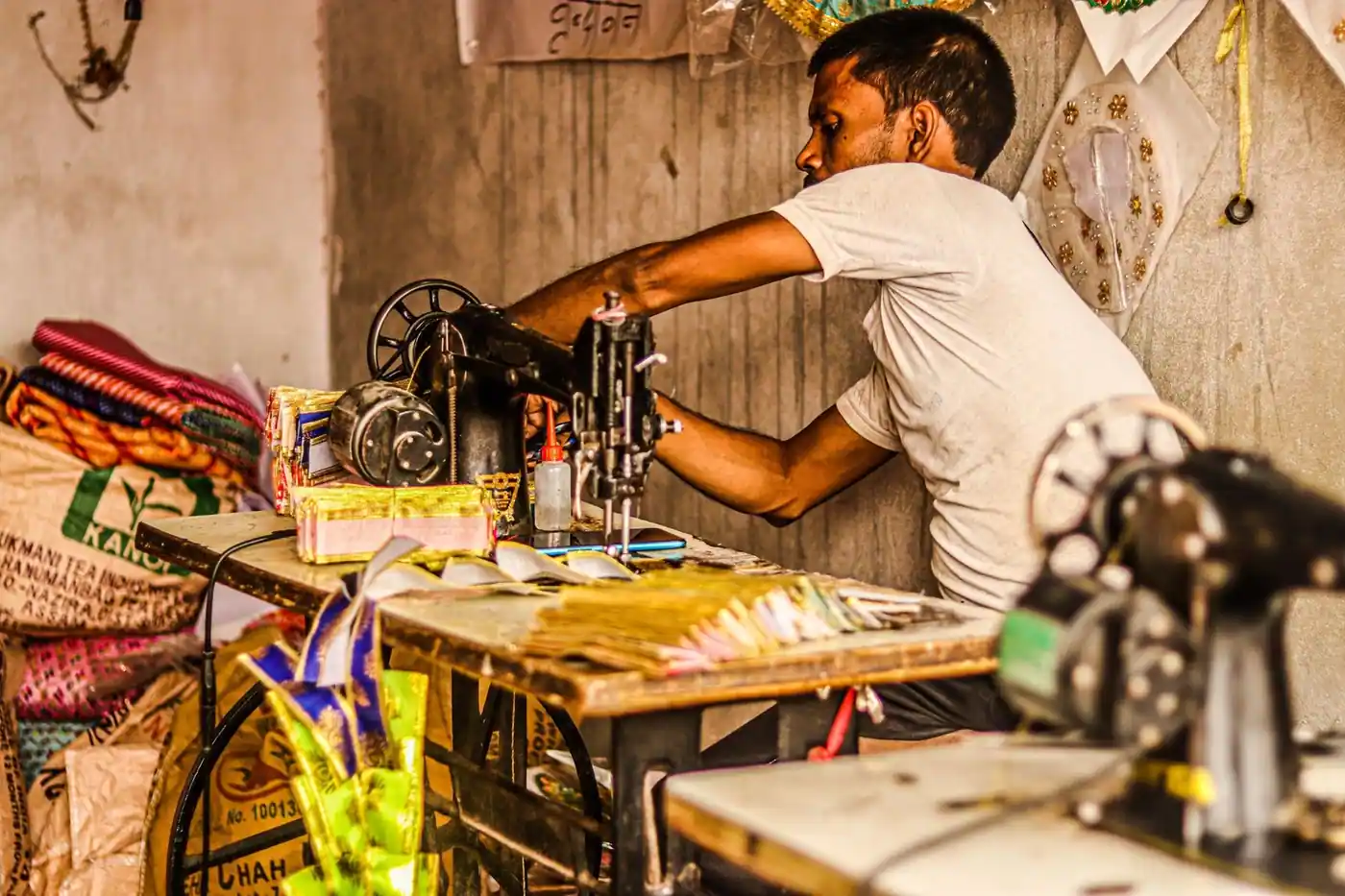Tailor in workshop using sewing machine; vibrant textiles surround him.