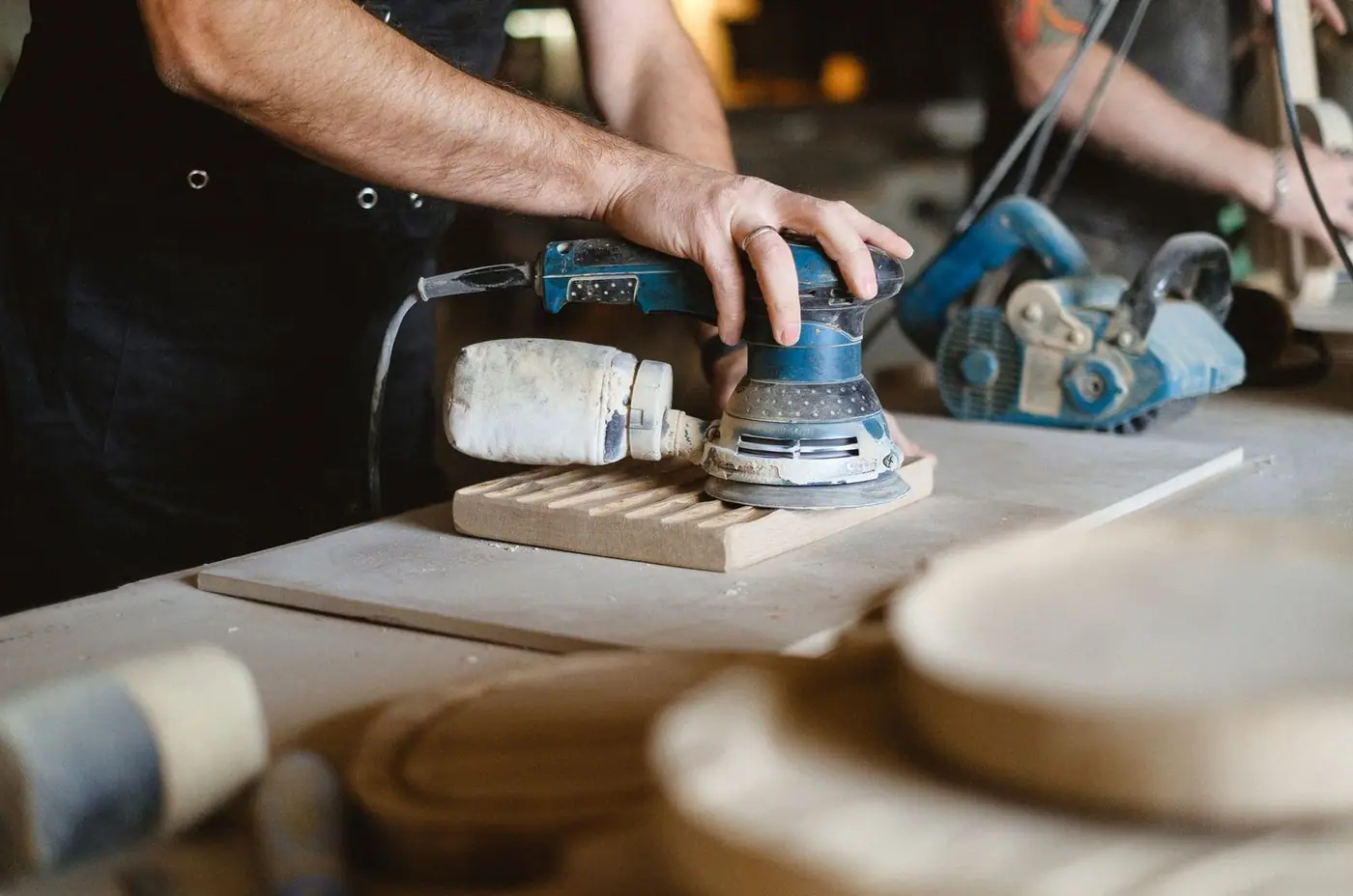 Unrecognizable professional male carpenter using random orbital sander while polishing wooden board during work at workbench in joinery stud