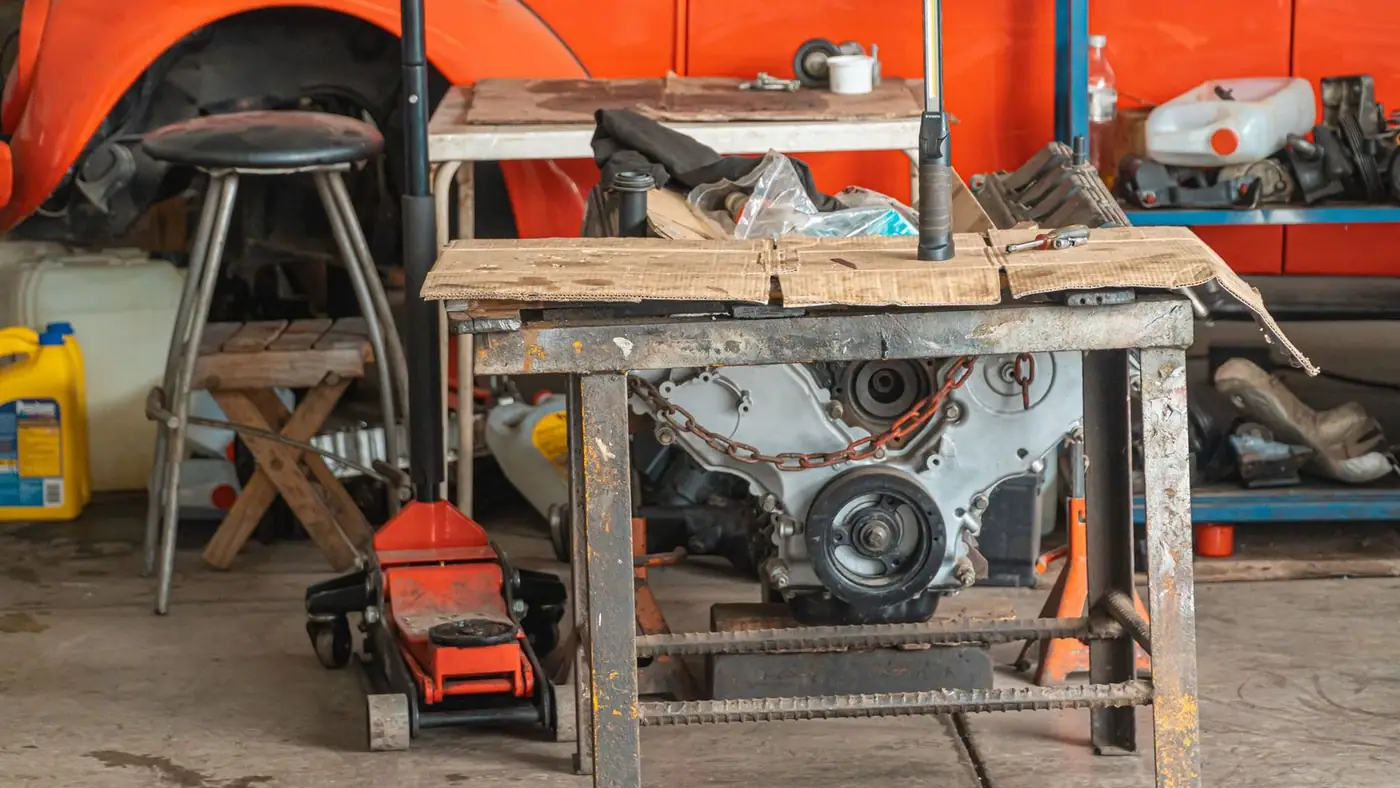 Old car engine on a workshop table with tools and a red car in the background.