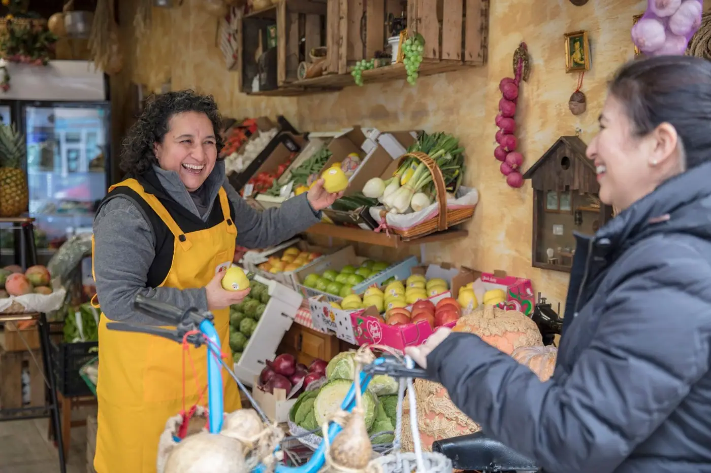 Two women smiling and exchanging fresh produce at a greengrocer's market.