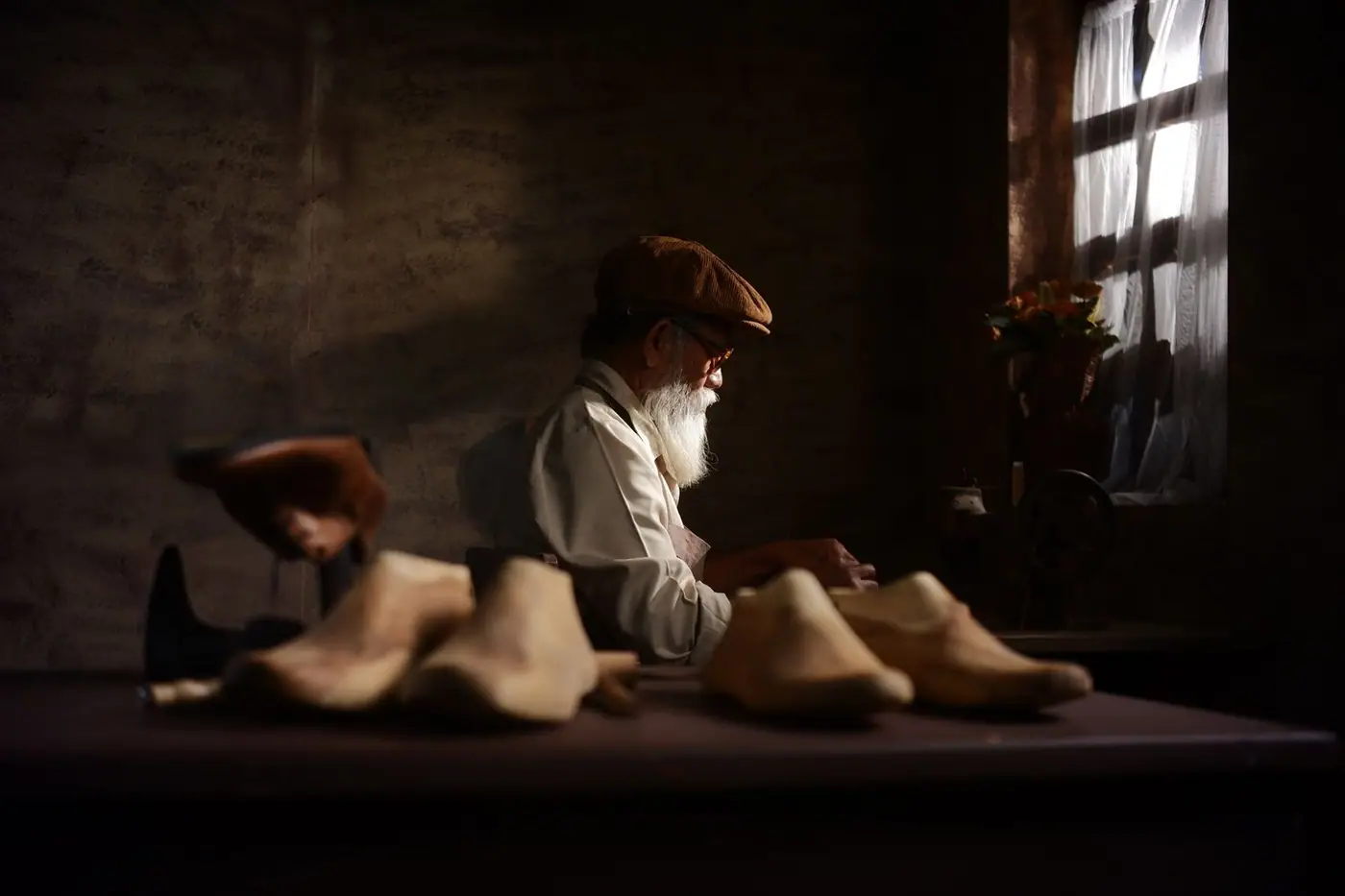 An elderly craftsman working with wooden tools at a bench in a dimly lit vintage workshop.