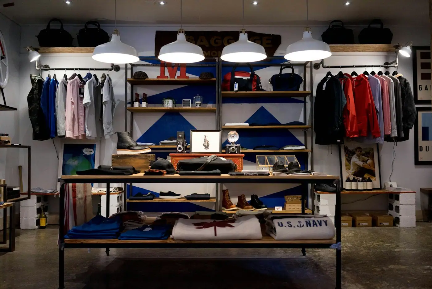Interior of a menswear boutique with pendant lights over shelves of bags, hanging shirts and jackets, and a central display table