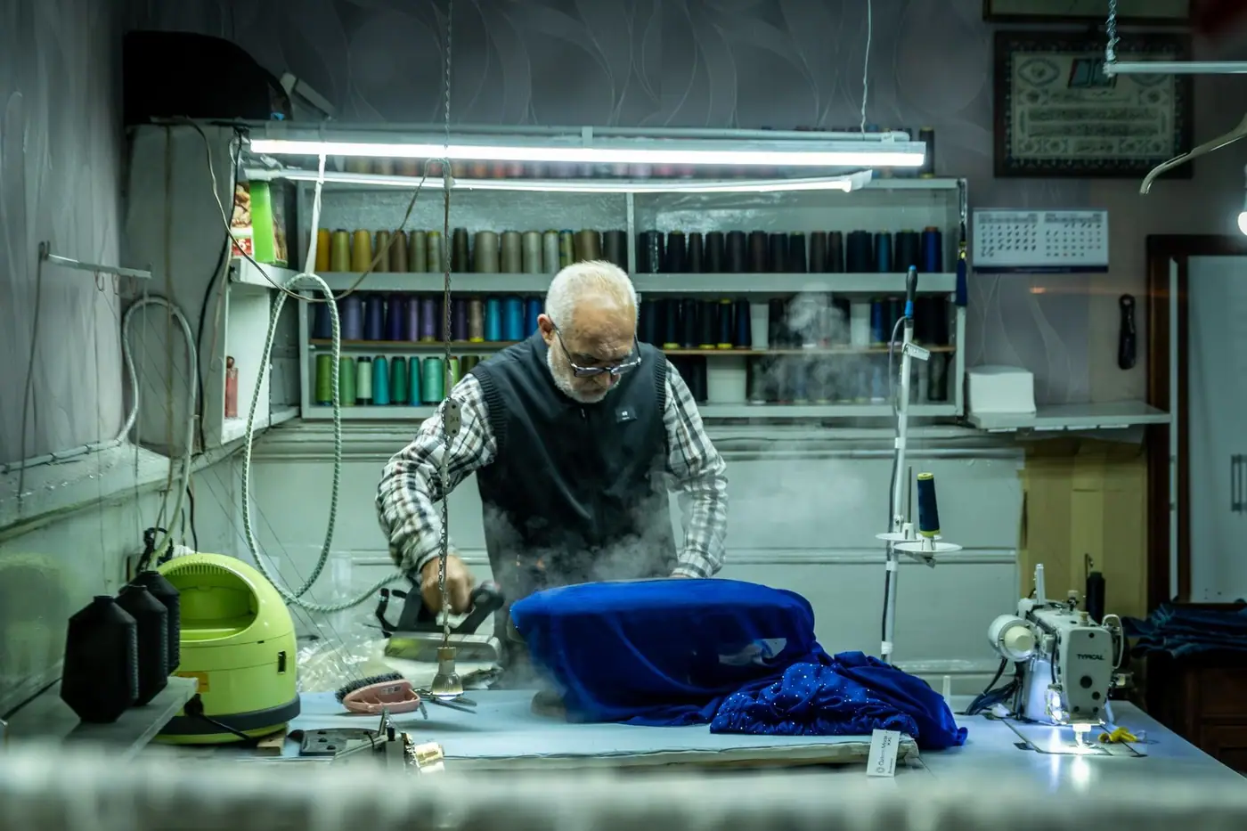 A tailor ironing blue fabric in a traditional sewing workshop surrounded by sewing tools