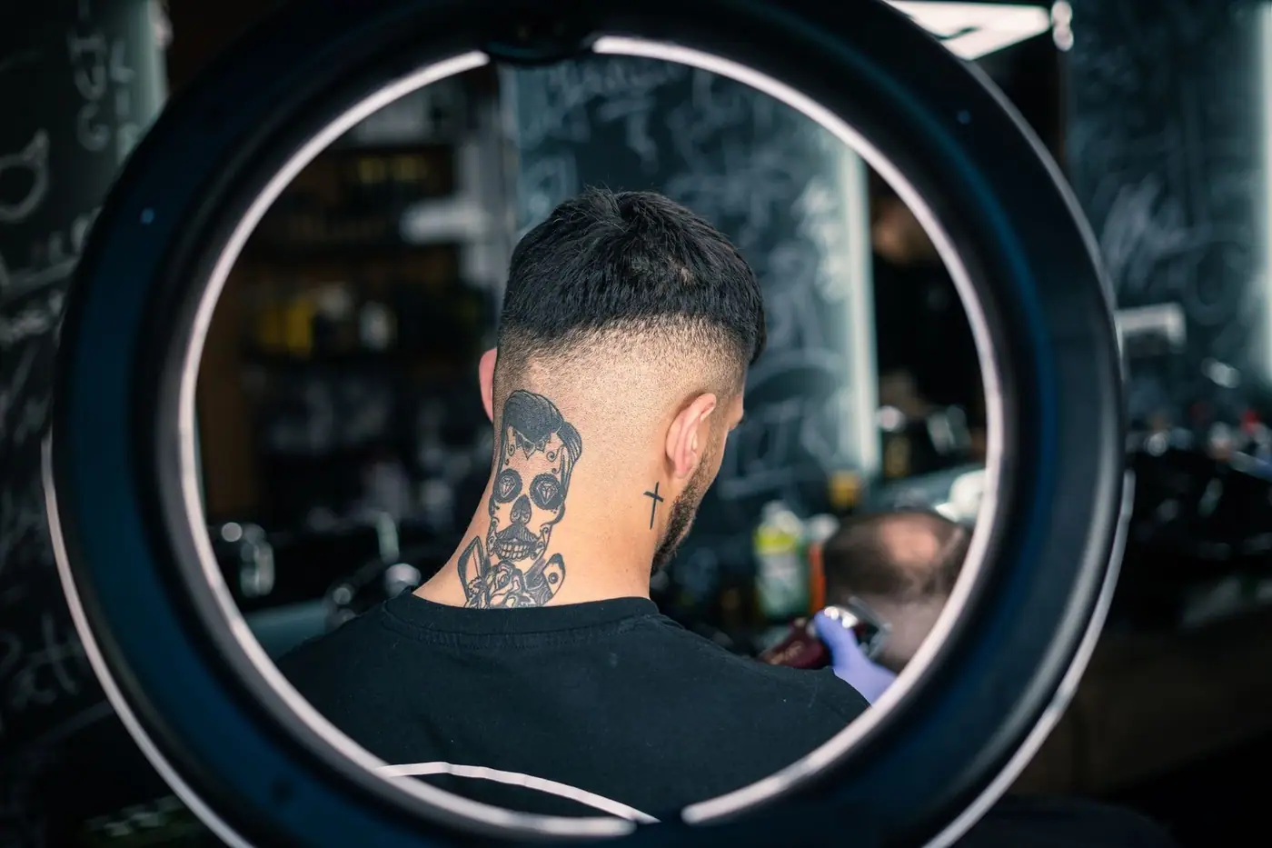 Back view of a man with a skull neck tattoo getting a haircut, lit by a ring light in a barbershop