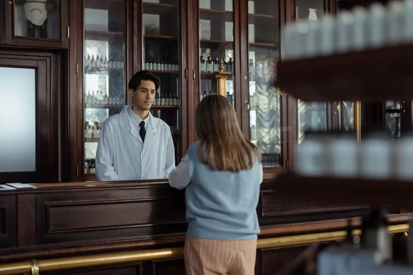 A pharmacist assists a customer at a vintage wooden pharmacy counter