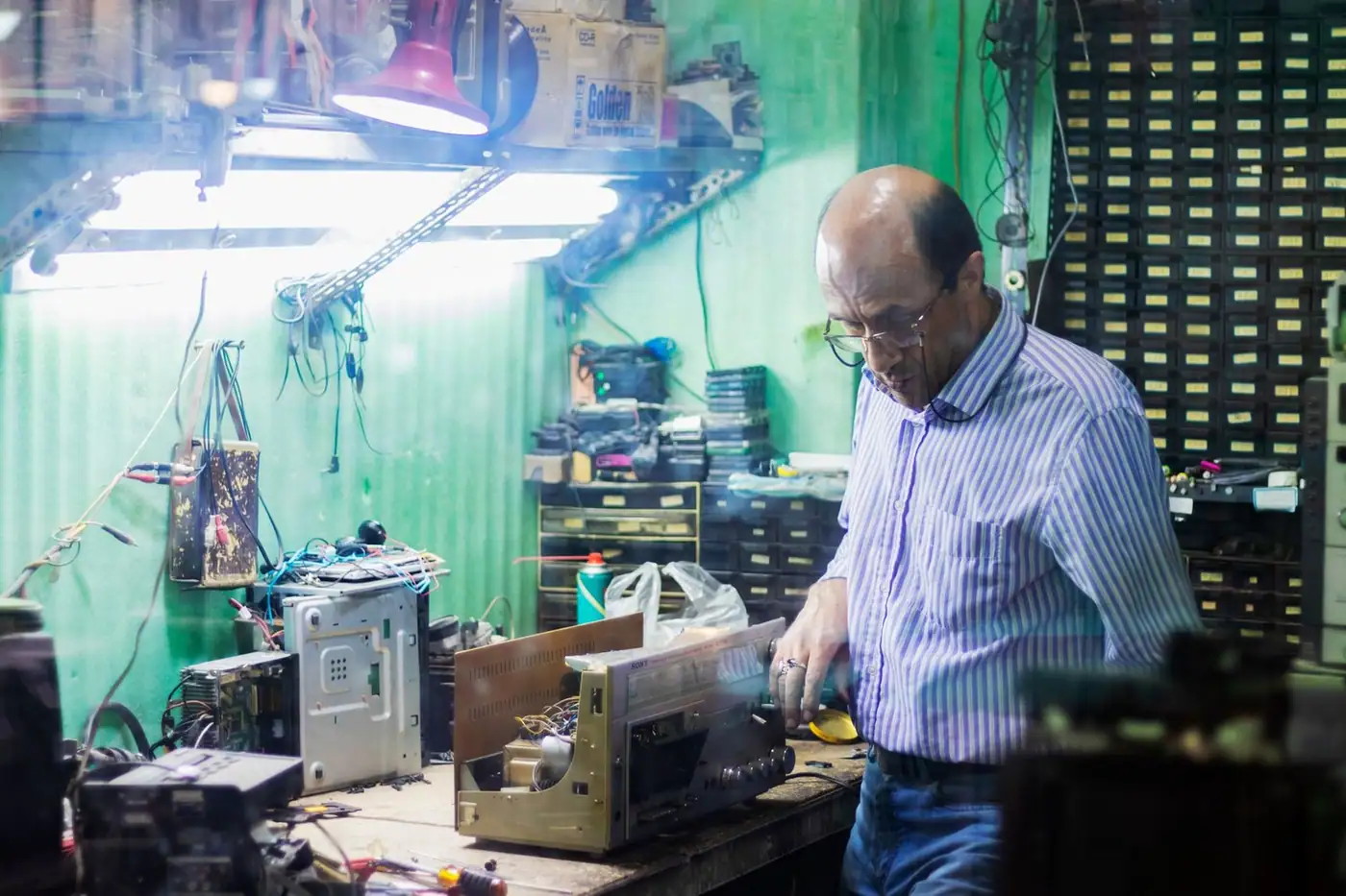 A technician bent over electronic equipment at a cluttered workshop bench full of tools
