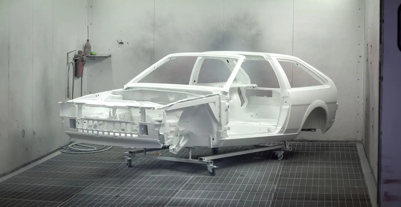 A vintage car frame being painted white in an industrial automotive paint booth