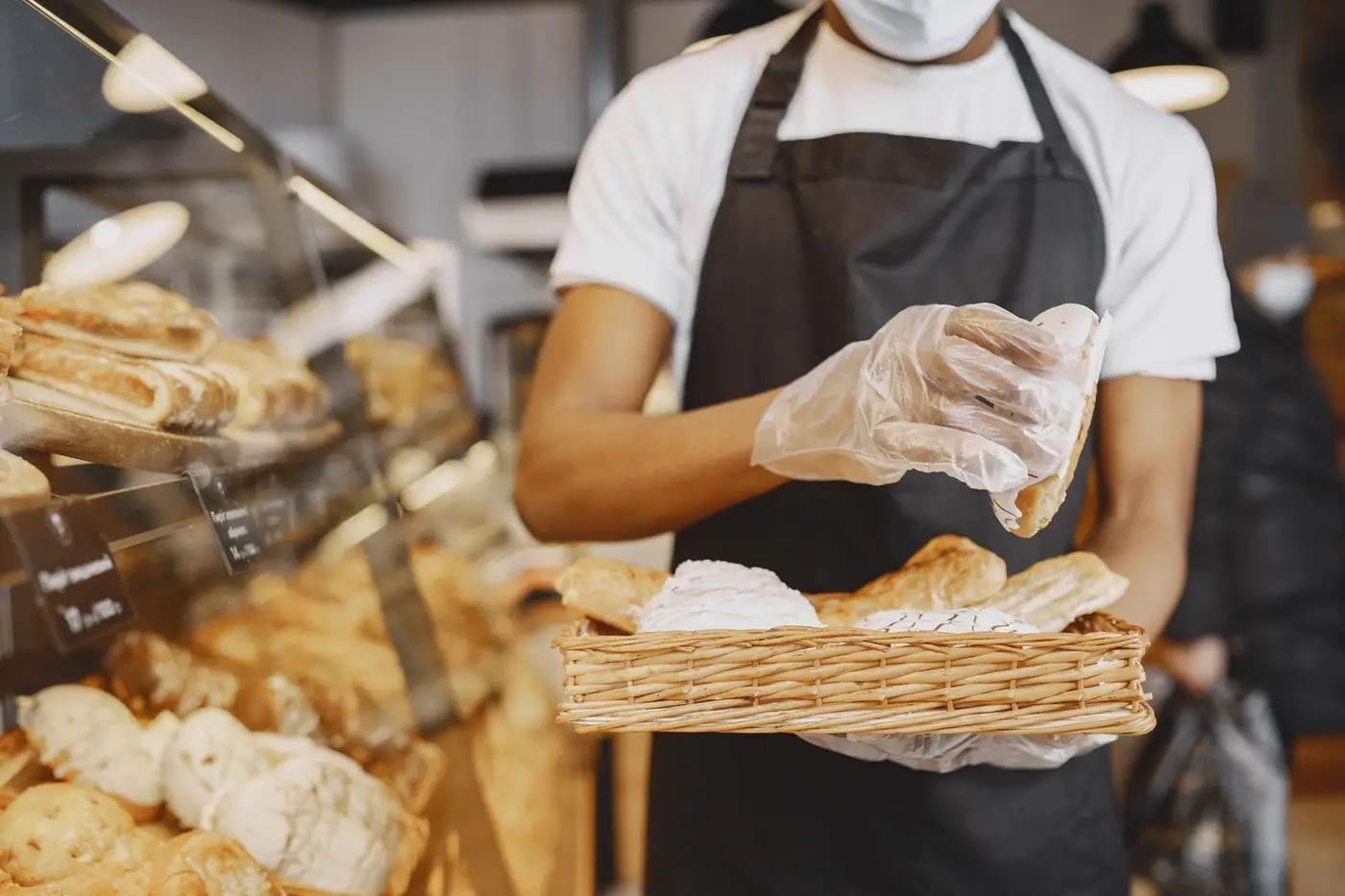 A baker wearing a mask and gloves prepares fresh pastries in a bakery