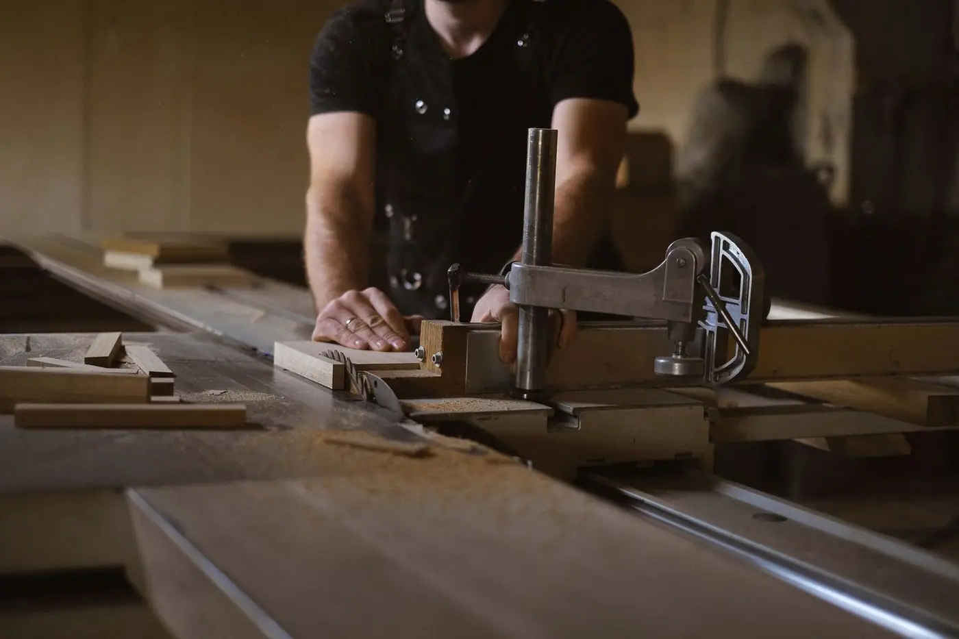 A woodworker in an apron at a sawbench beside a stack of wood