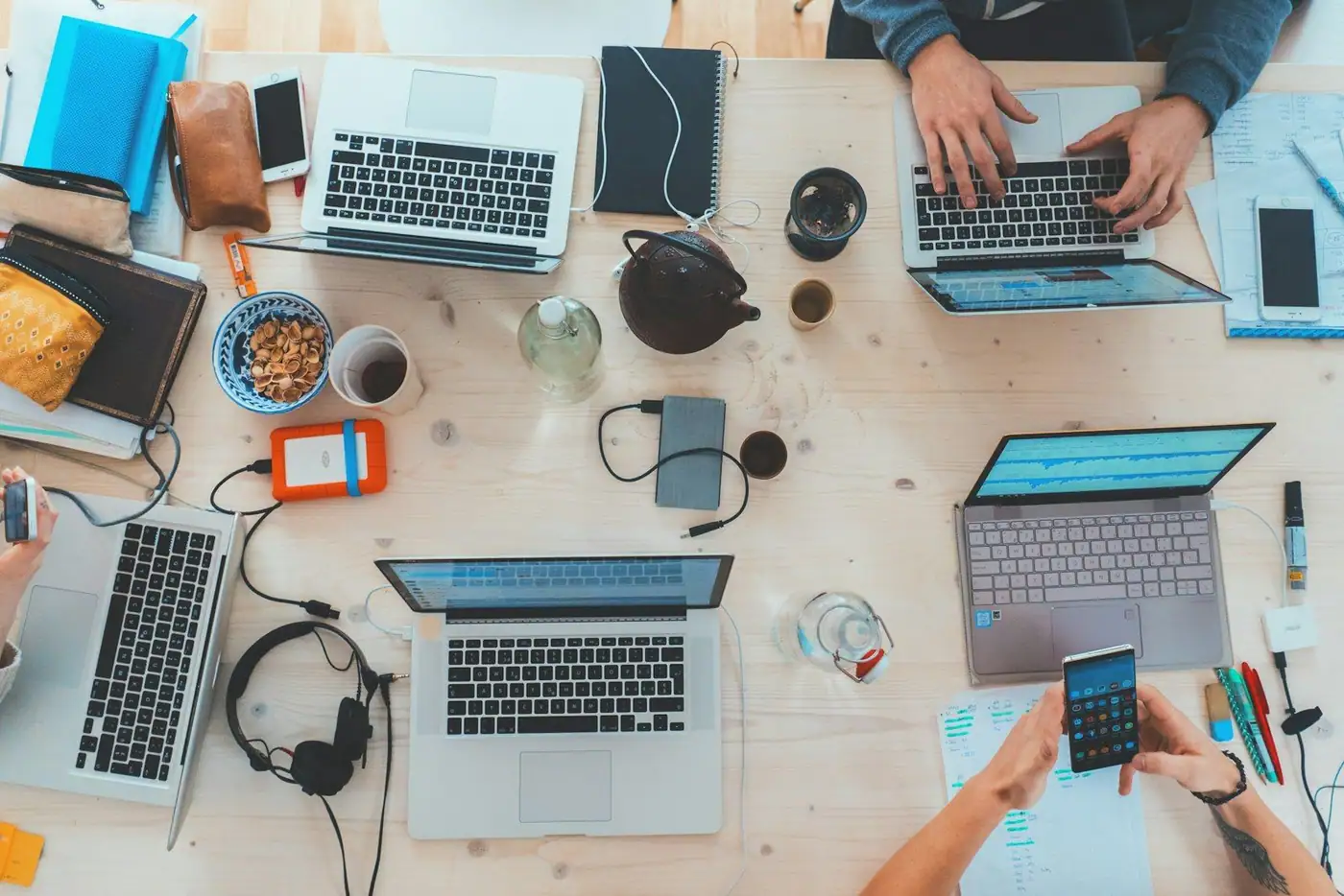 Overhead view of a busy wooden table with laptops, phones, notebooks and drinks