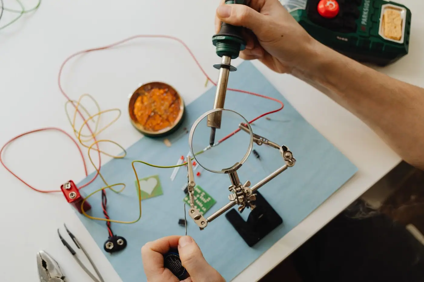 Hands soldering a circuit board using a magnifying glass and tools, seen from above