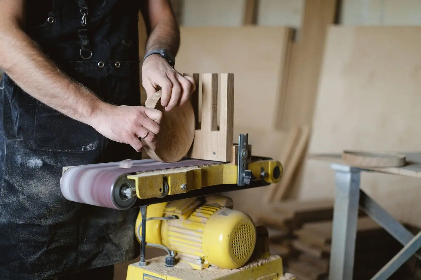 A tradesman using a belt and disc sander to shape a piece of wood, with a stack of timber behind