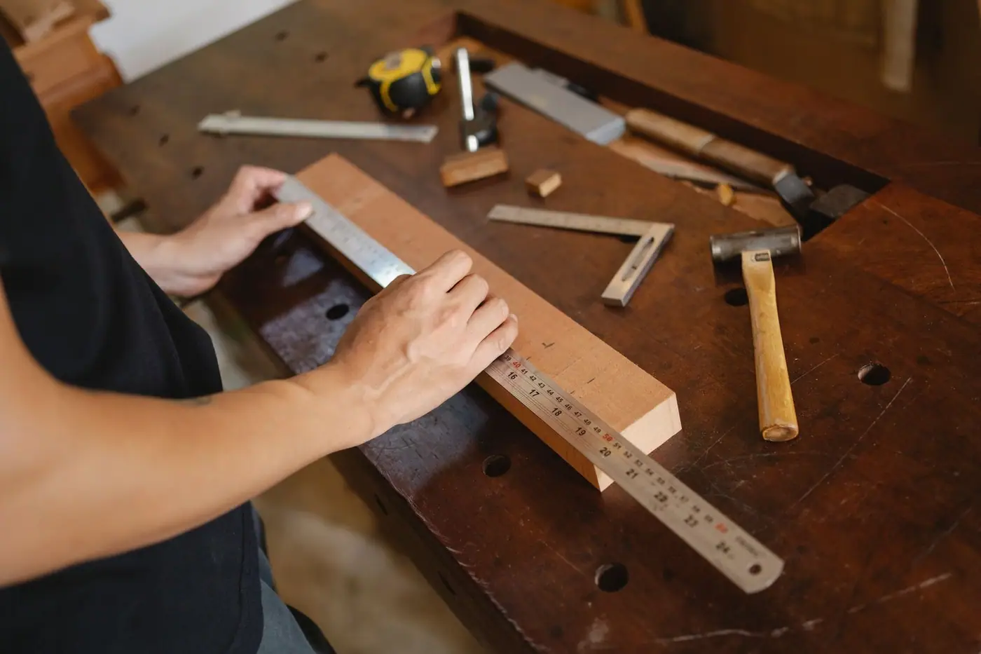 Hands measuring a timber plank on a table with a hammer nearby