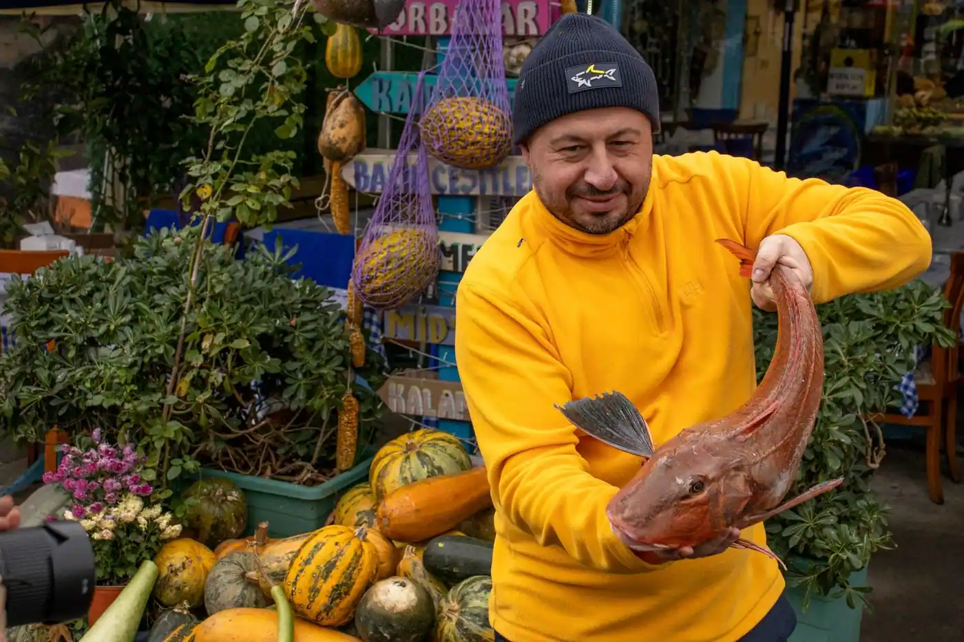 A man in a yellow jacket holding a fish at an outdoor market with various produce
