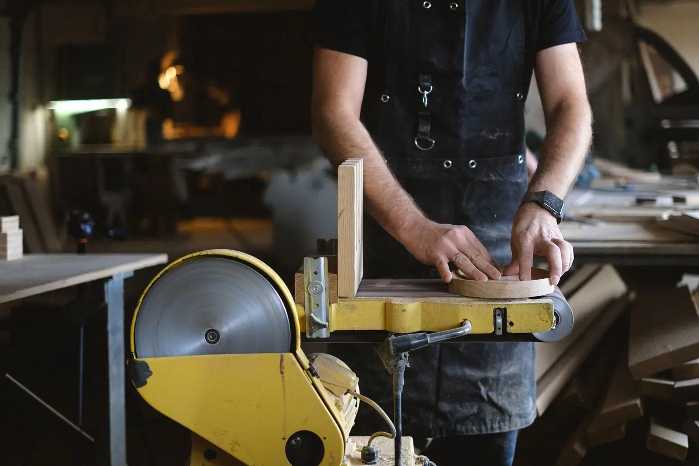 A craftsman in an apron shaping a round piece of wood on a belt and disc sander