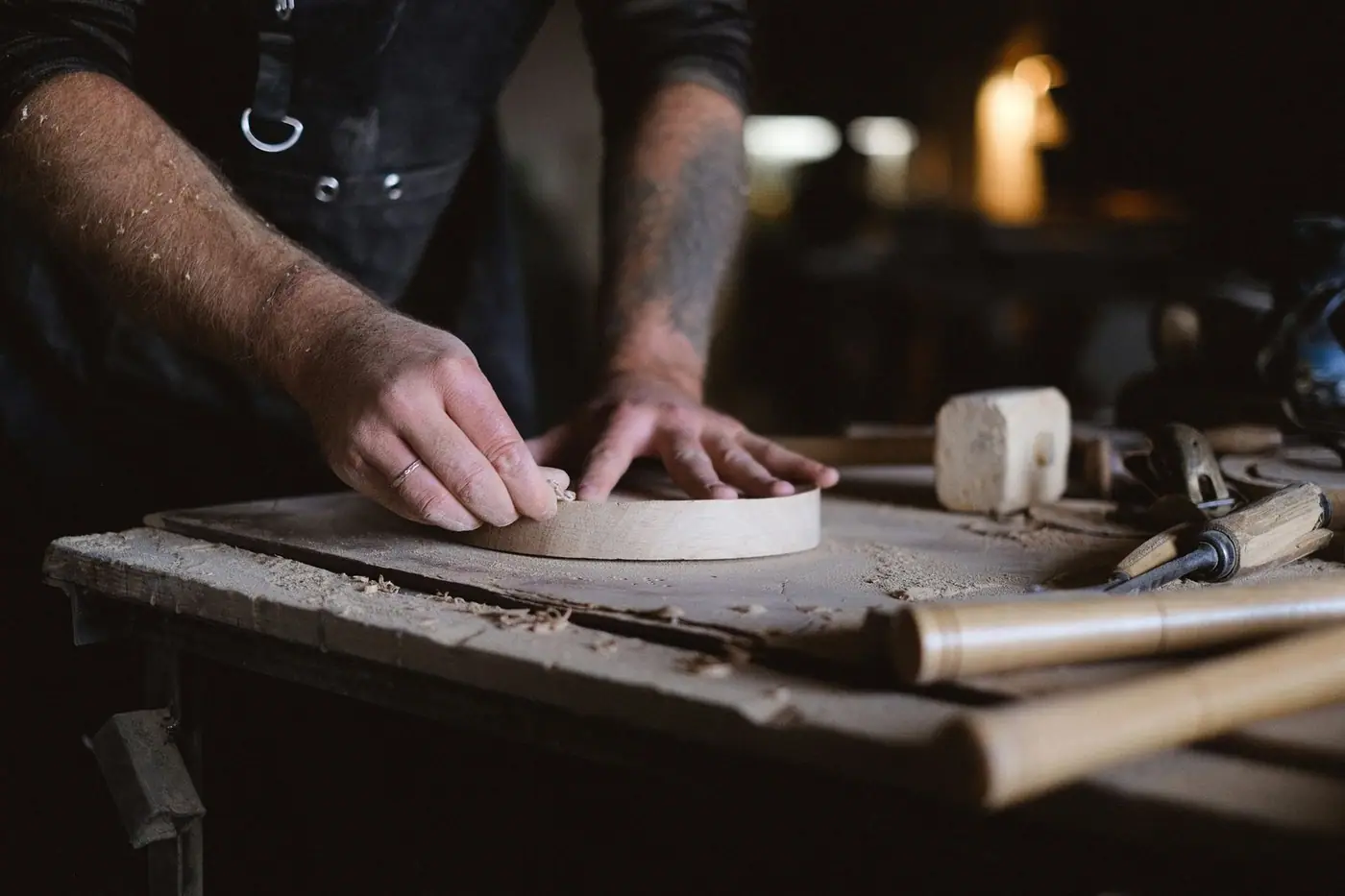A craftsman sanding a round wooden piece at a workbench surrounded by hand tools