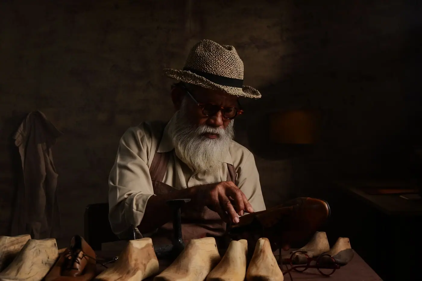 An elderly craftsman working carefully at his bench in a dimly lit workshop