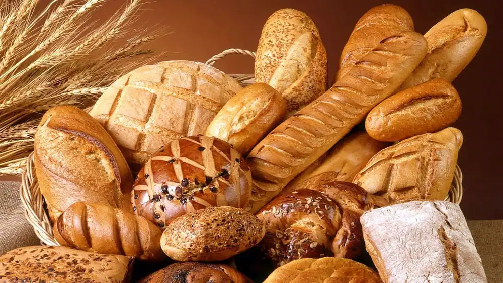 Assortment of breads, baguettes and rolls piled in a wicker basket with wheat stalks against a warm brown background.