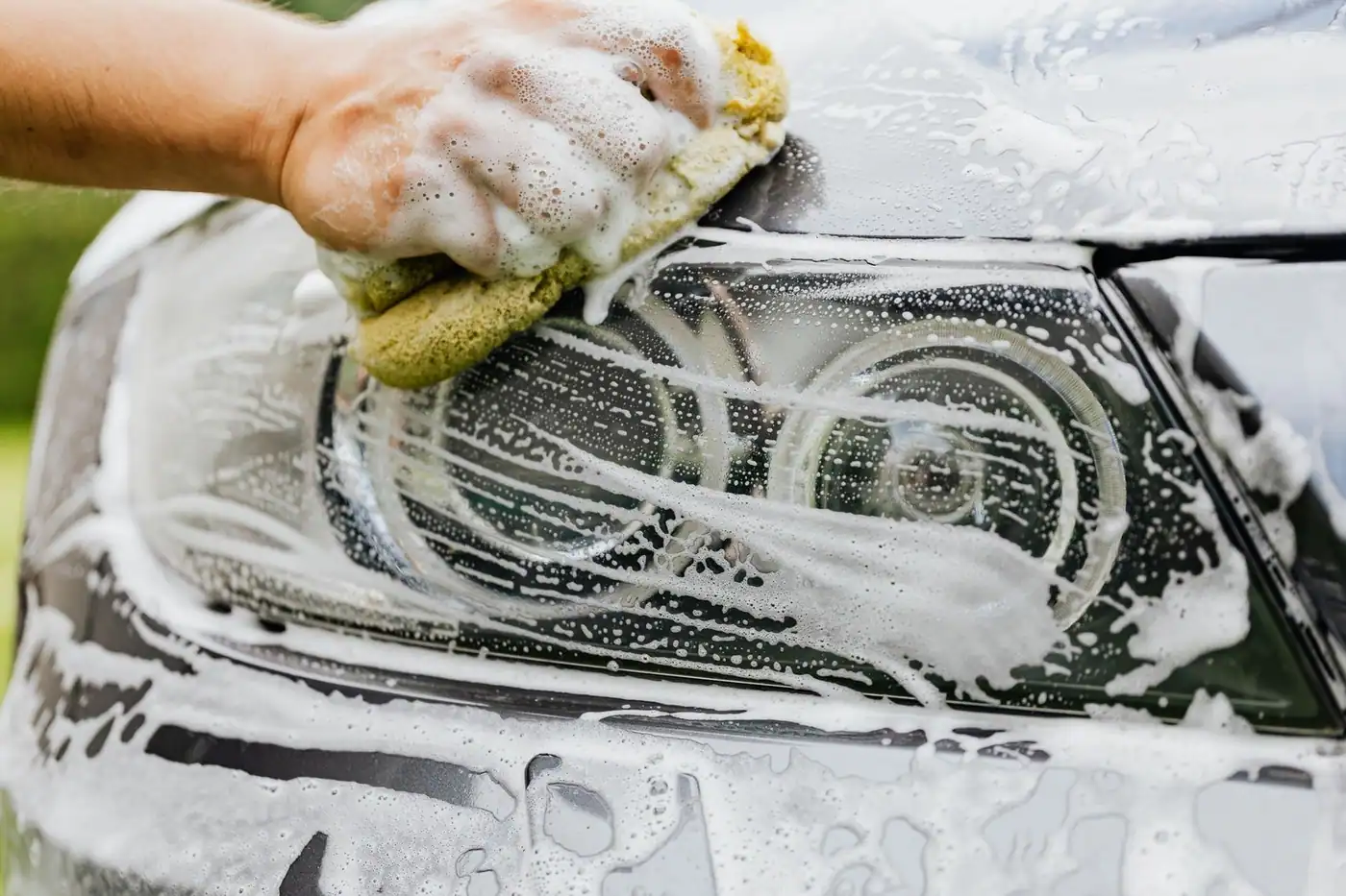 A hand scrubbing a car headlight with a sponge and soap suds