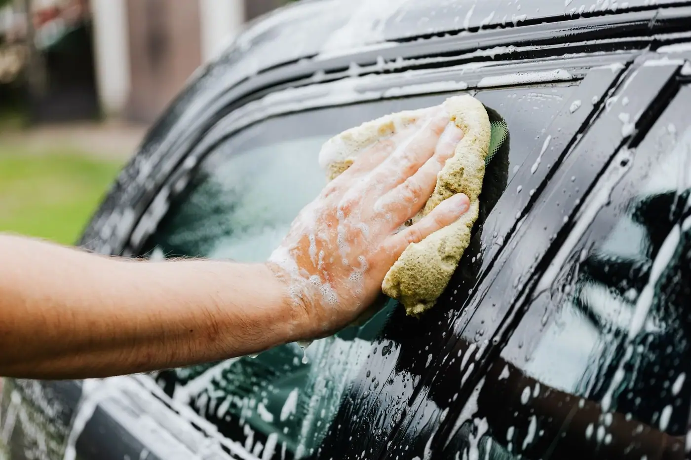 A hand scrubbing a car window with a sponge covered in soap suds