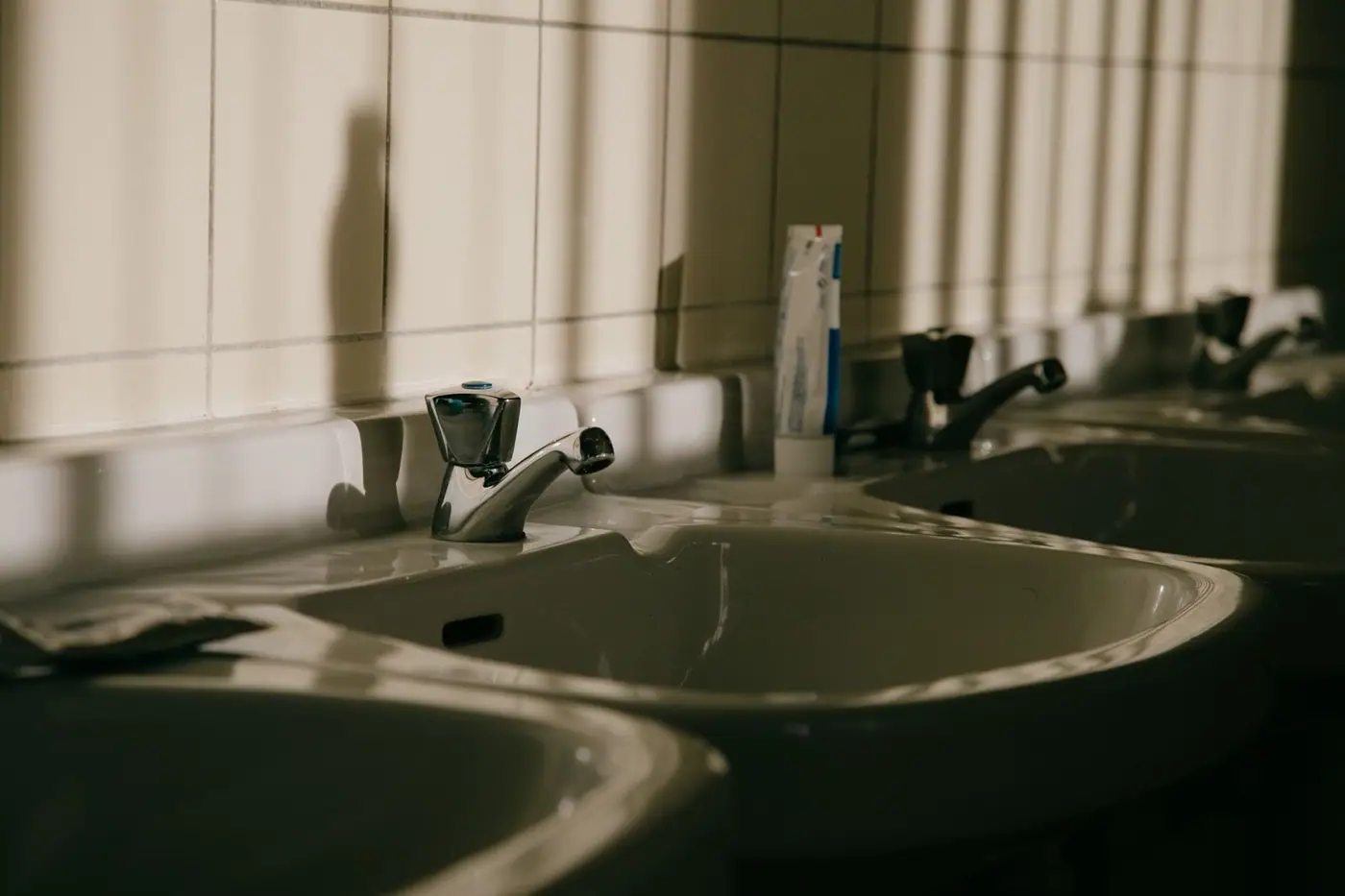 A row of bathroom sinks with taps and shadows on tiled walls