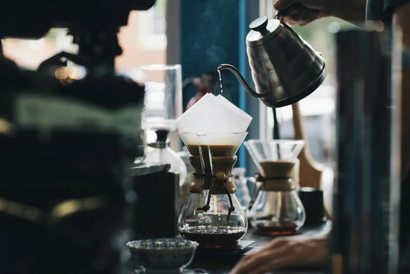 A hand pours hot water from a gooseneck kettle into a Chemex pour-over coffee maker by a window