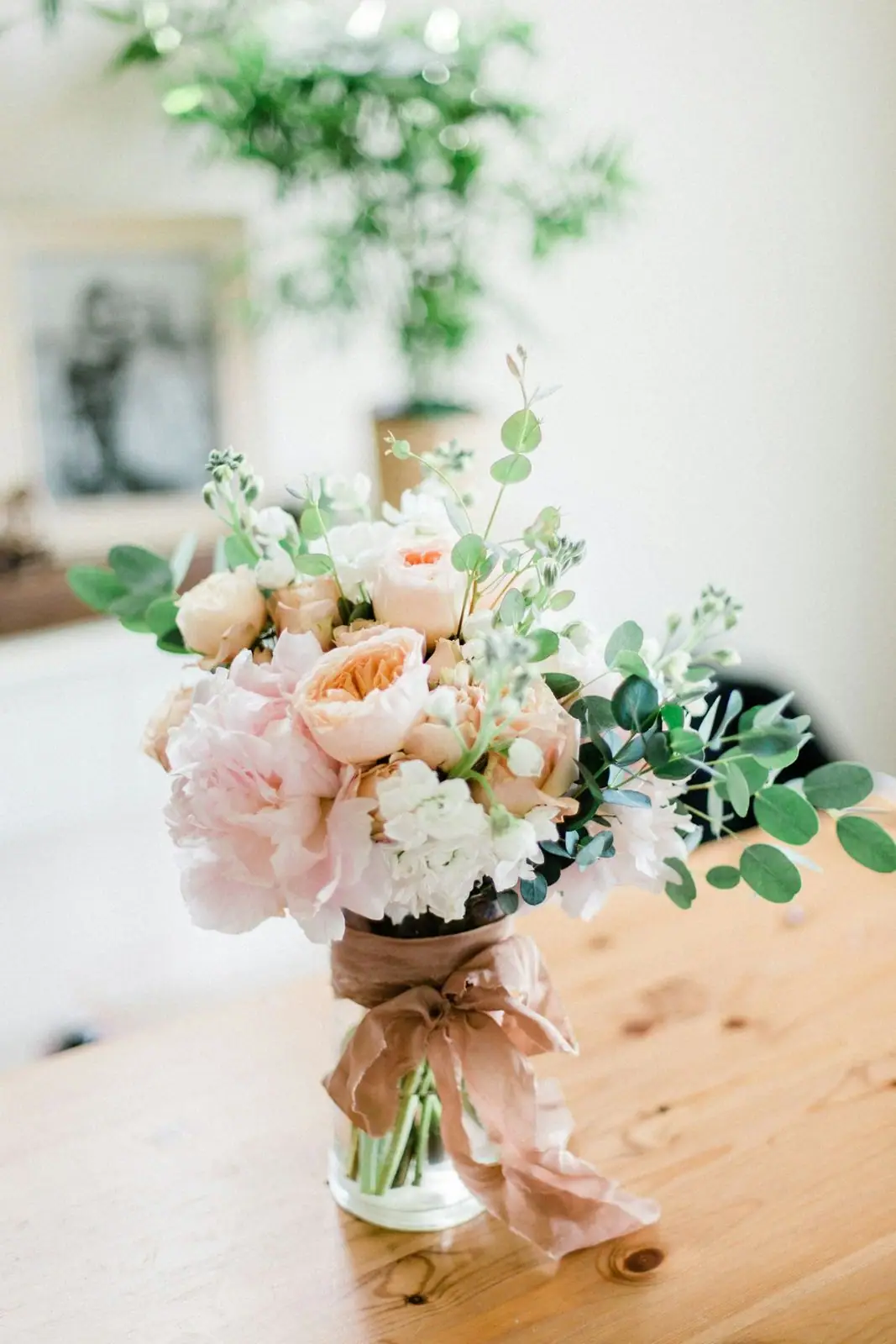 A soft, romantic bouquet of blush peonies, peach garden roses and eucalyptus tied with a satin ribbon, set in a glass jar on a wooden table