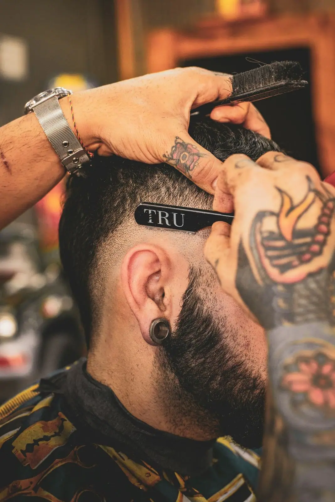 Close-up of a barber using a straight razor labeled 'TRU' and a brush to shape a client's fade haircut and sideburns.