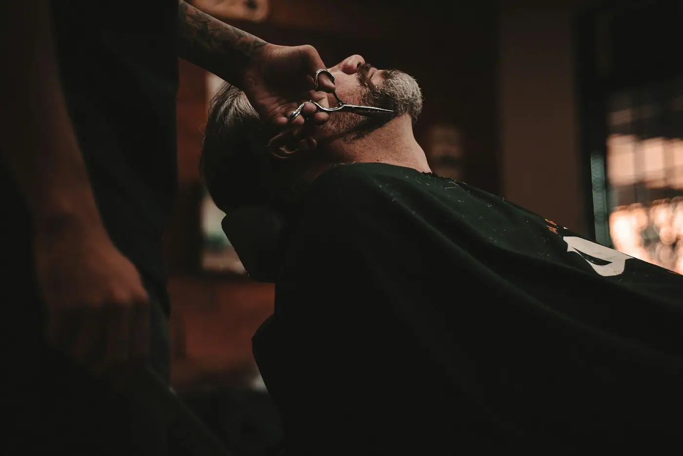 A barber trimming a reclining customer's beard with scissors under warm, low lighting