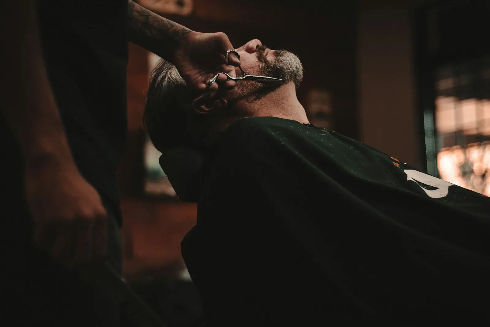 A barber with a tattooed arm trims the beard of a reclining bearded man using scissors, in dim warm lighting.