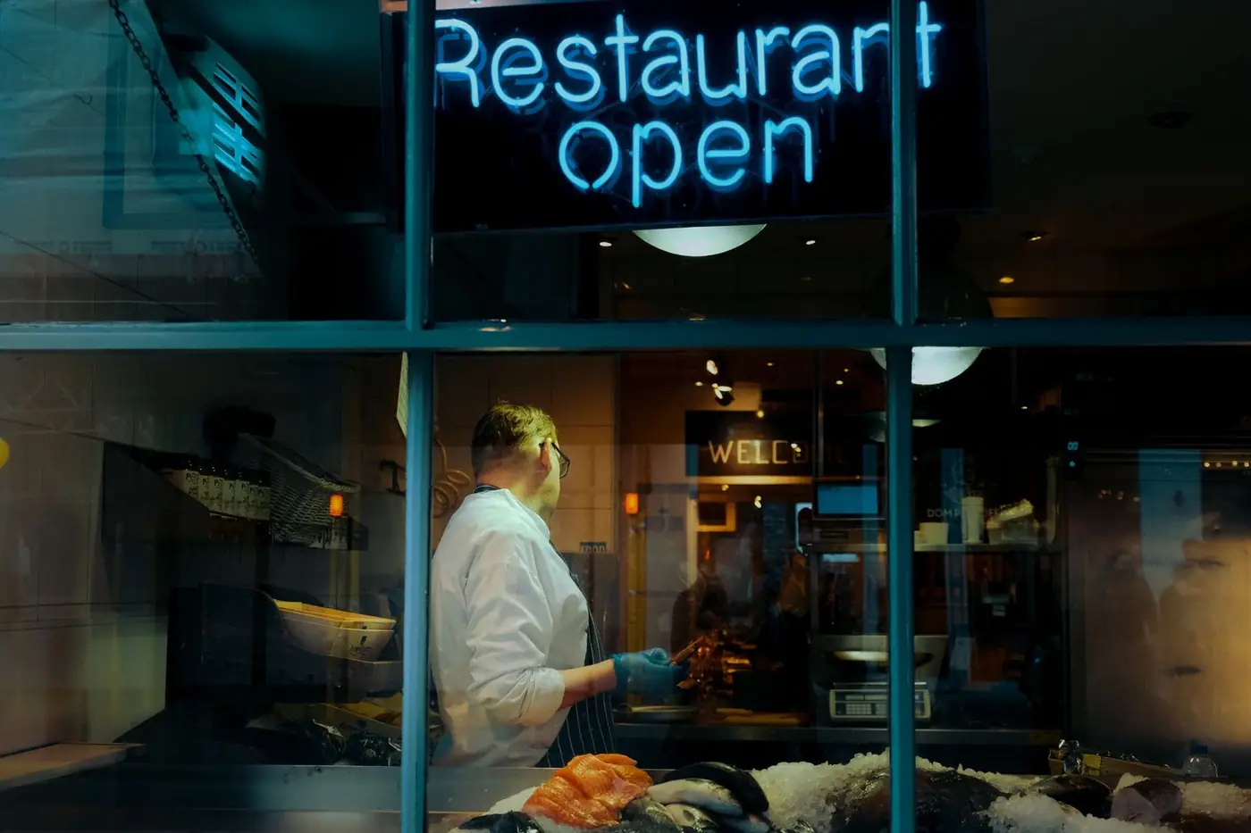 A chef at work behind a window with a neon 'Restaurant Open' sign