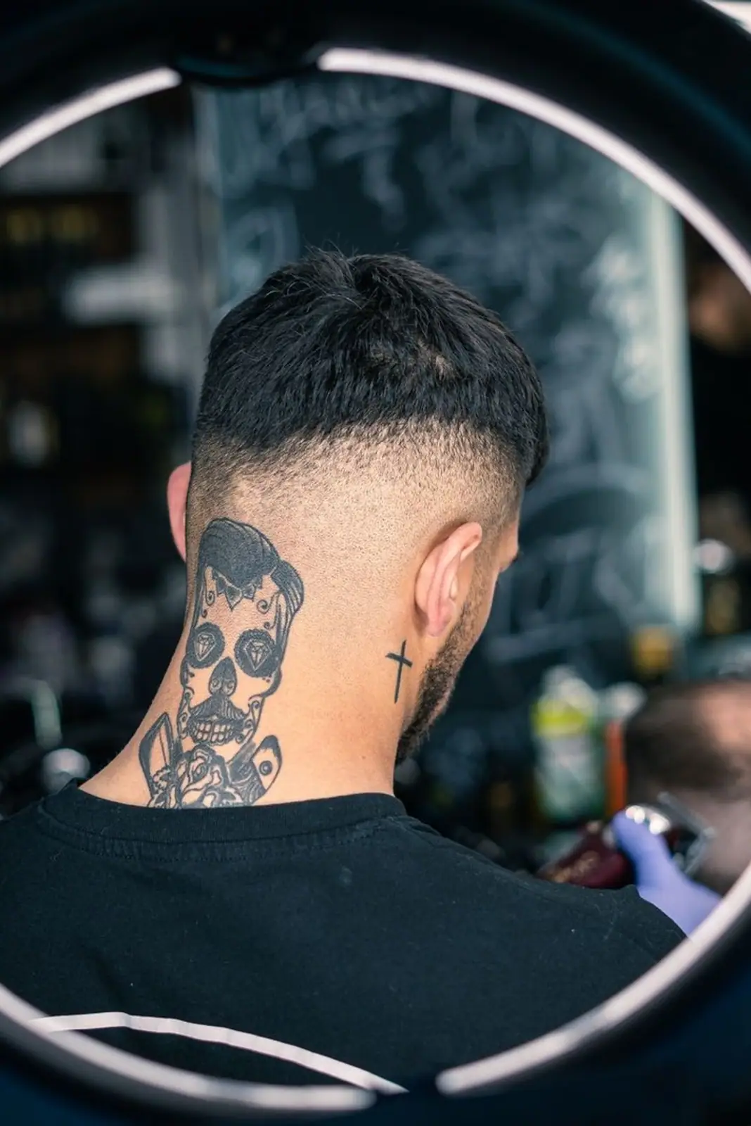 Back view of man with skull neck tattoo getting a haircut surrounded by a ring light in a barbershop.