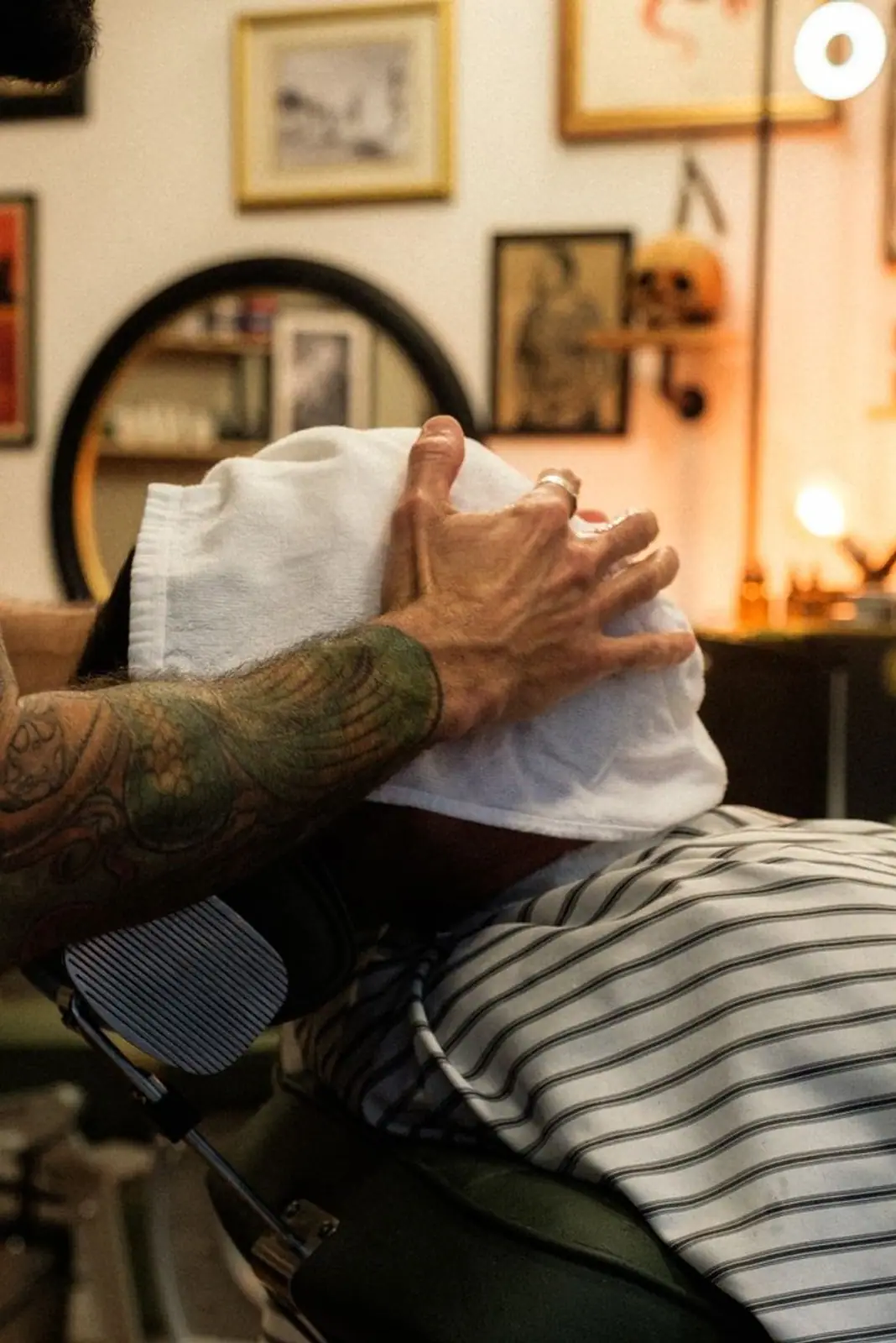 A barber with tattoo sleeves applies a hot towel in a vintage-style barbershop setting.